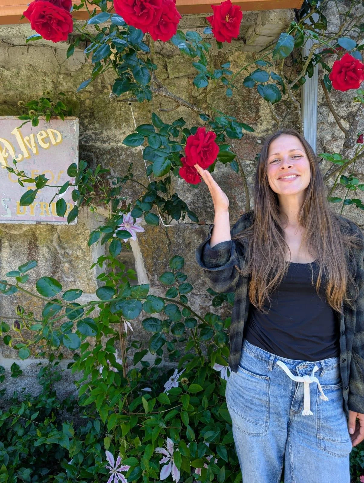 A young woman with long brown hair smiling at the camera, standing next to a flowering bush with large red roses and white flowers, in front of a stone wall with a faded sign.