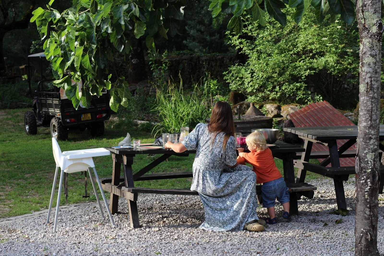 Eine Frau sitzt an einem Holztisch im Garten mit einem jungen Kind, beide scheinen zu essen oder zu trinken. Es ist Nachmittag, und die Umgebung ist grün mit Bäumen, Gras und einer gekachelten Struktur im Hintergrund. Es befindet sich ein weißer Hoch