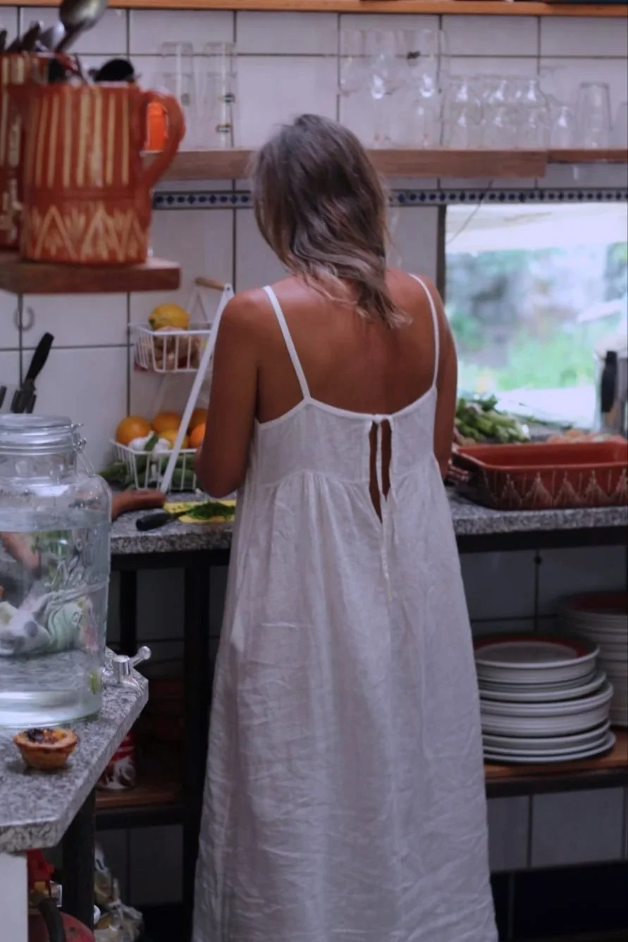 Frau in einem weißen Kleid beim Kochen in einer Küche mit weißen Fliesen und offenen Regalen.