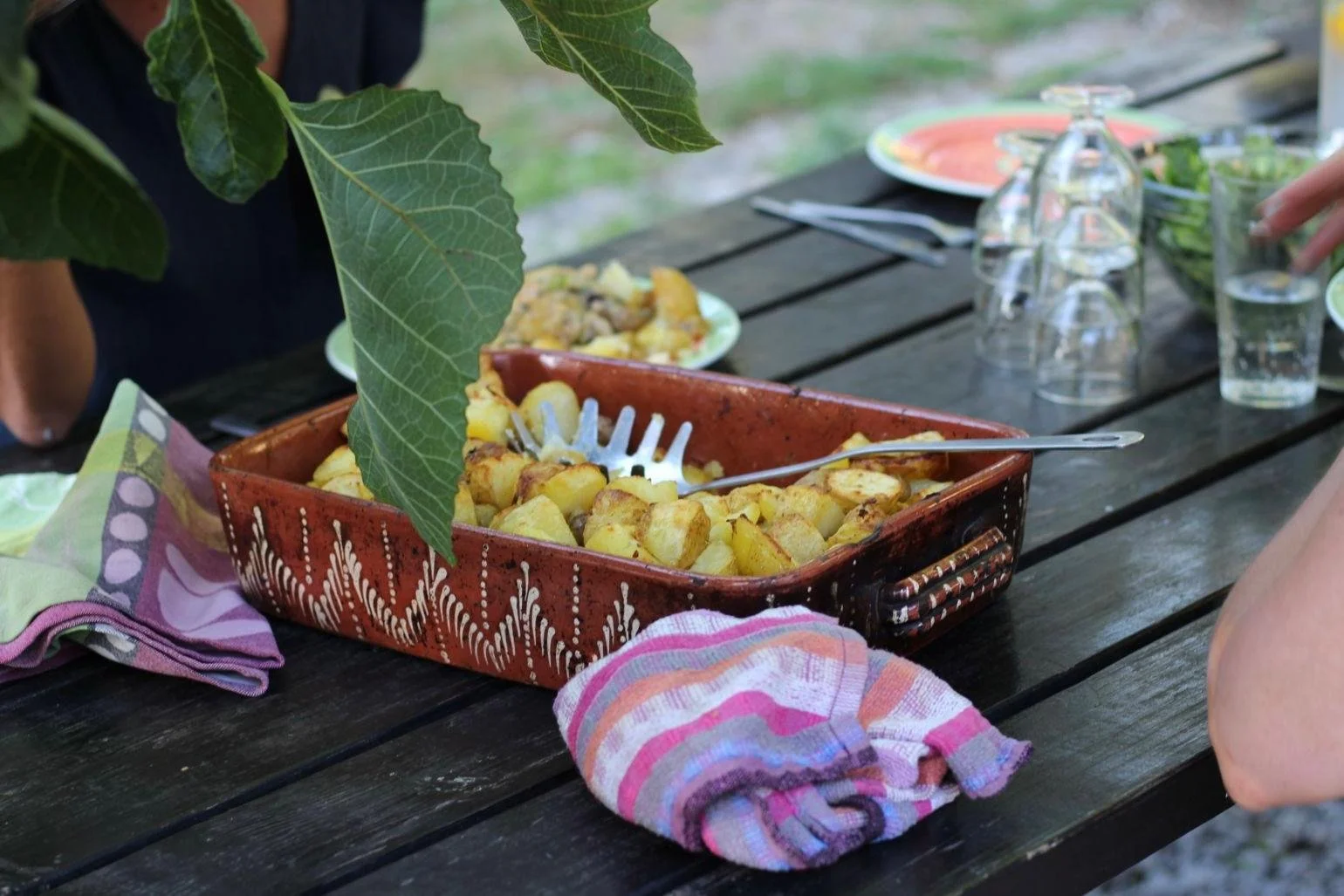 Ein Erdbeer-Dessert in einer roten, dekorierten Schale, auf einem dunklen Holztisch mit bunten Servietten und Gläsern, umgeben von Gartenblättern im Hintergrund.