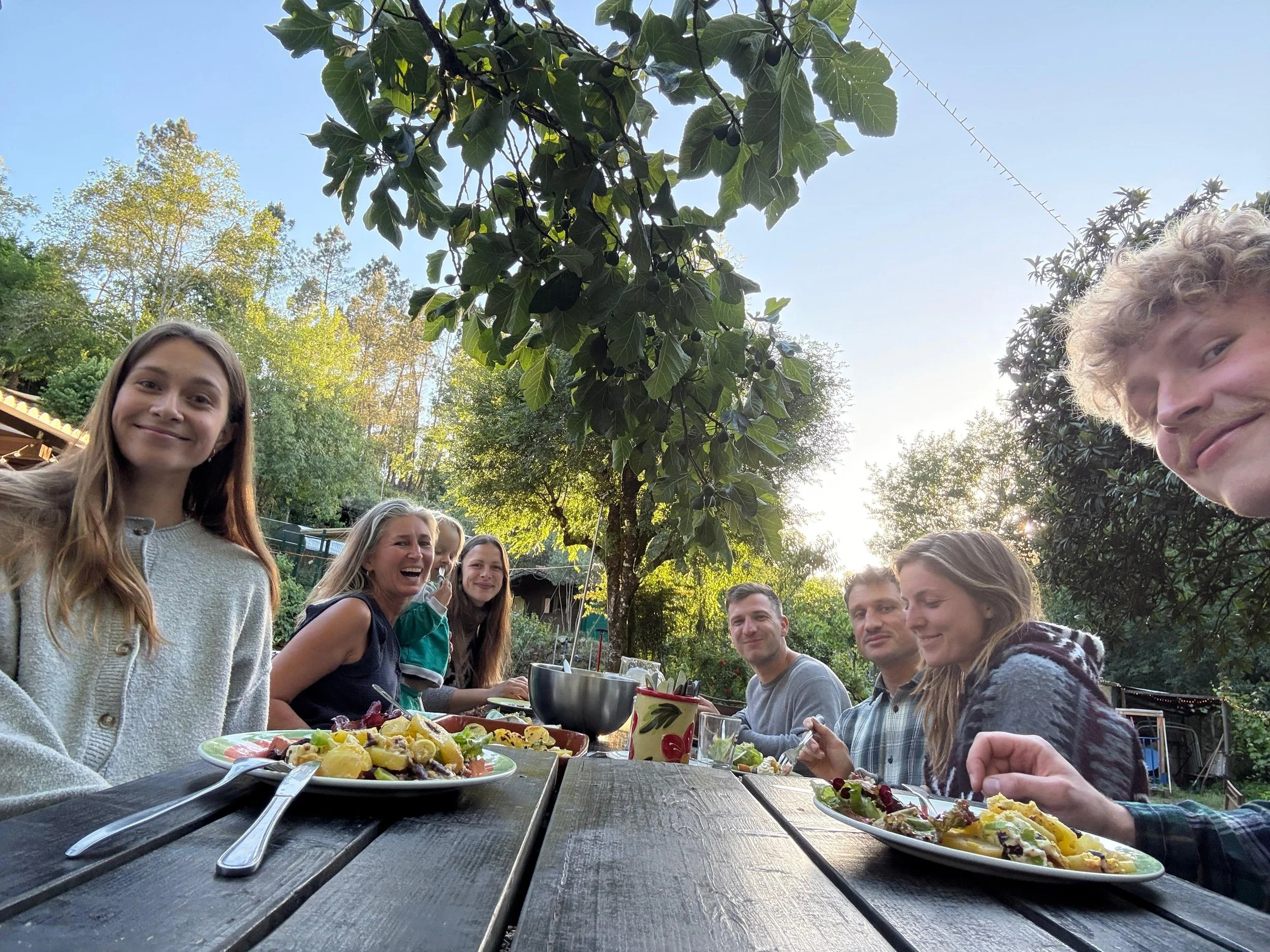 A group of people enjoying an outdoor dinner at a wooden table in a garden with trees and greenery, with plates of food and drinks visible.