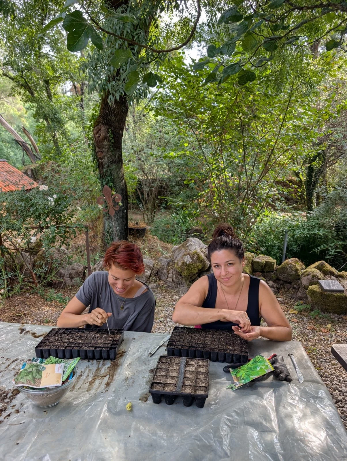Two women planting seeds in seed trays outdoors in a lush green garden.
