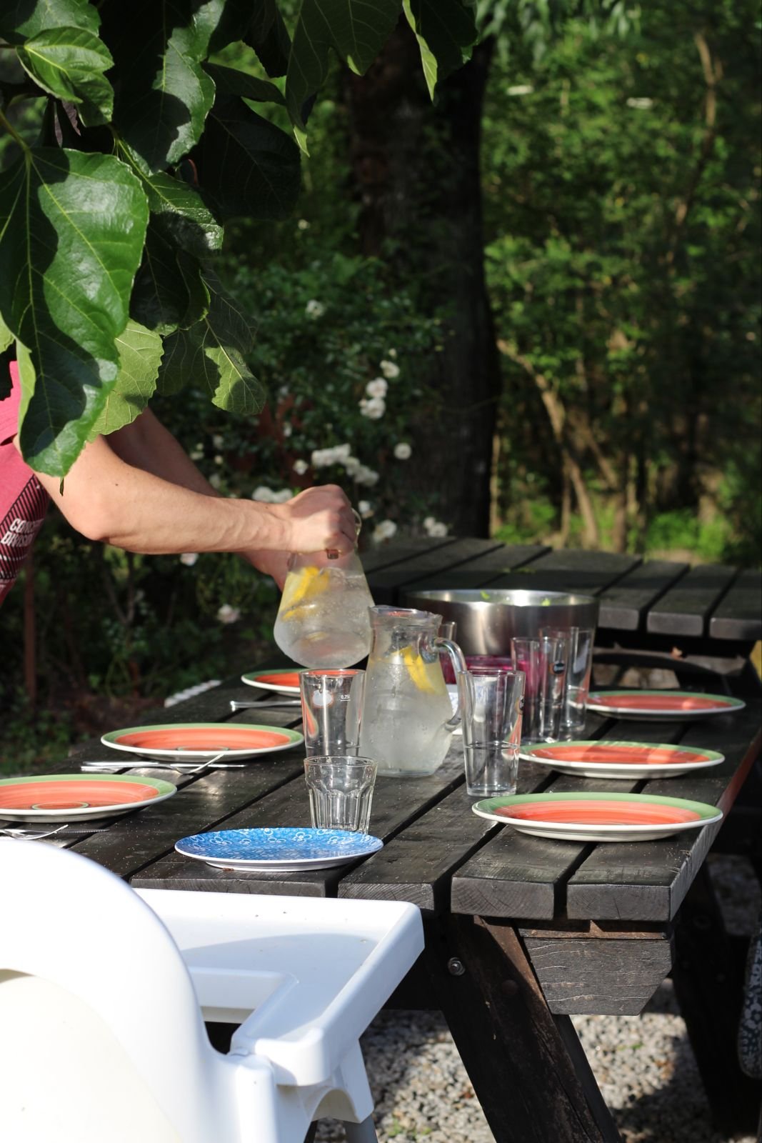 Person pouring lemonade from a pitcher into glasses on a wooden outdoor table, surrounded by greenery and plates for a summer meal.