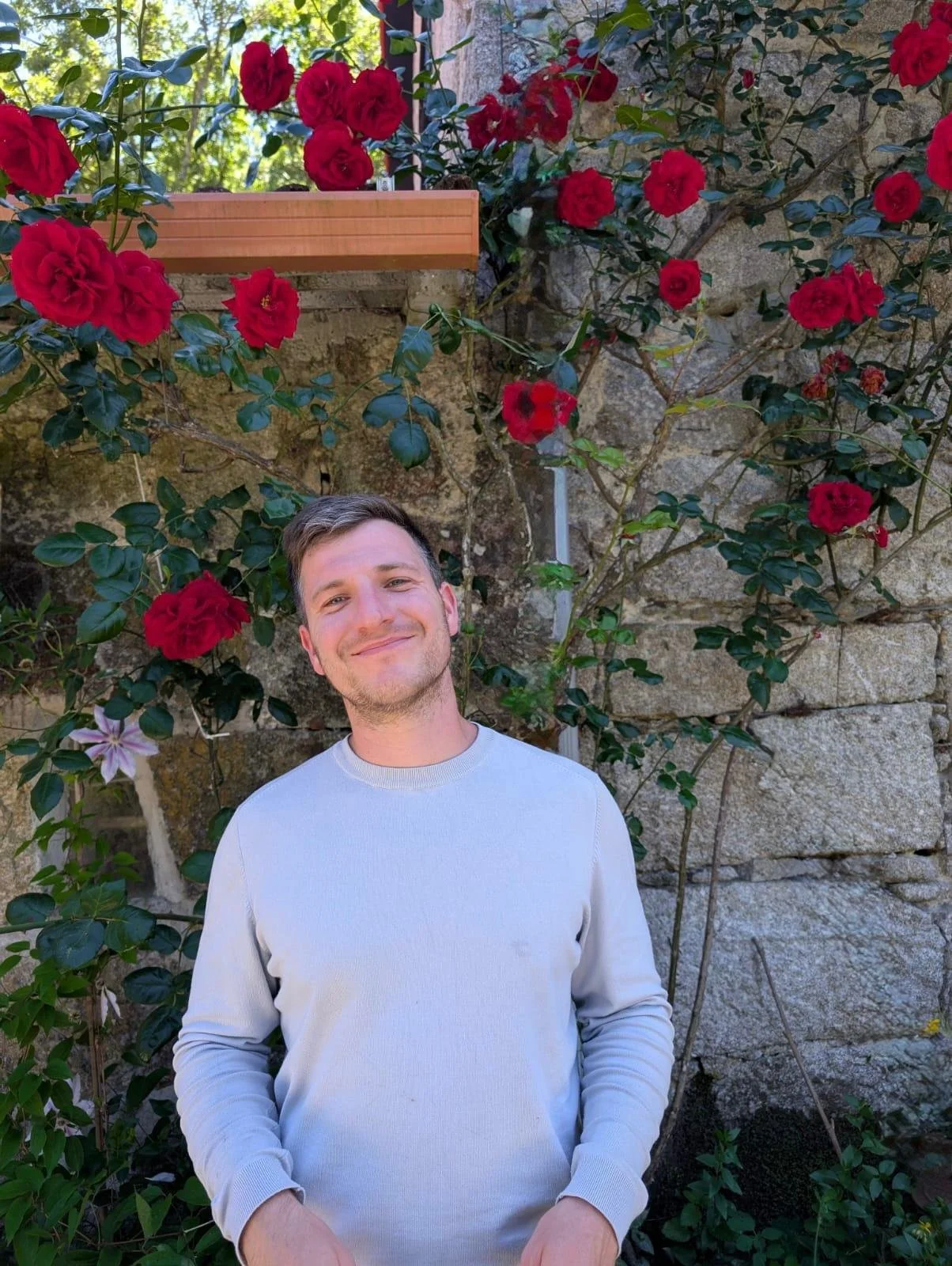 A man smiling in front of a wall with red roses and green foliage.