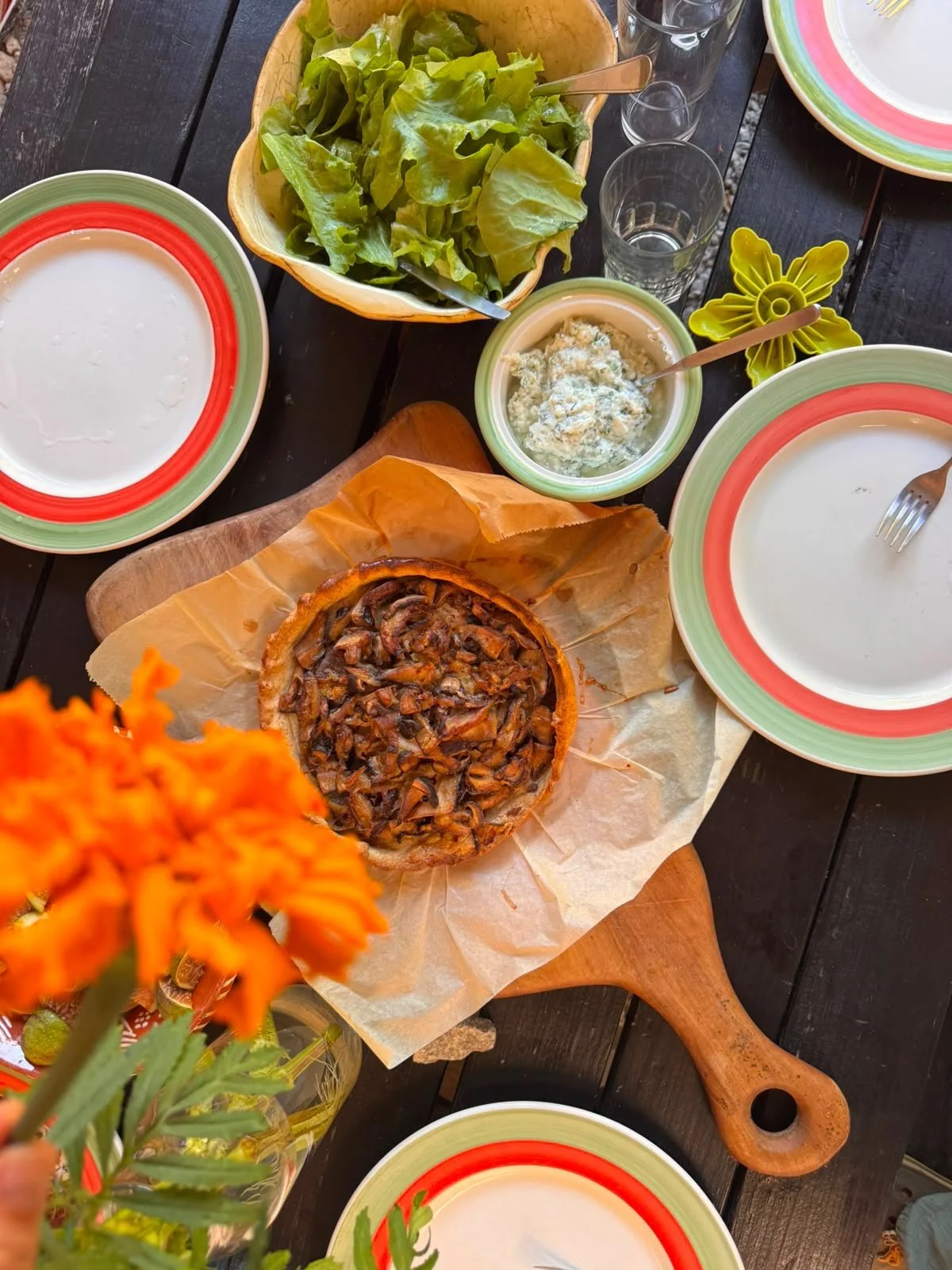 A table set with bowls of salad, tortilla pie, shredded cheese, and various dishes. There are glasses, plates, and a wooden serving paddle, with a flower in an orange vase in the foreground.