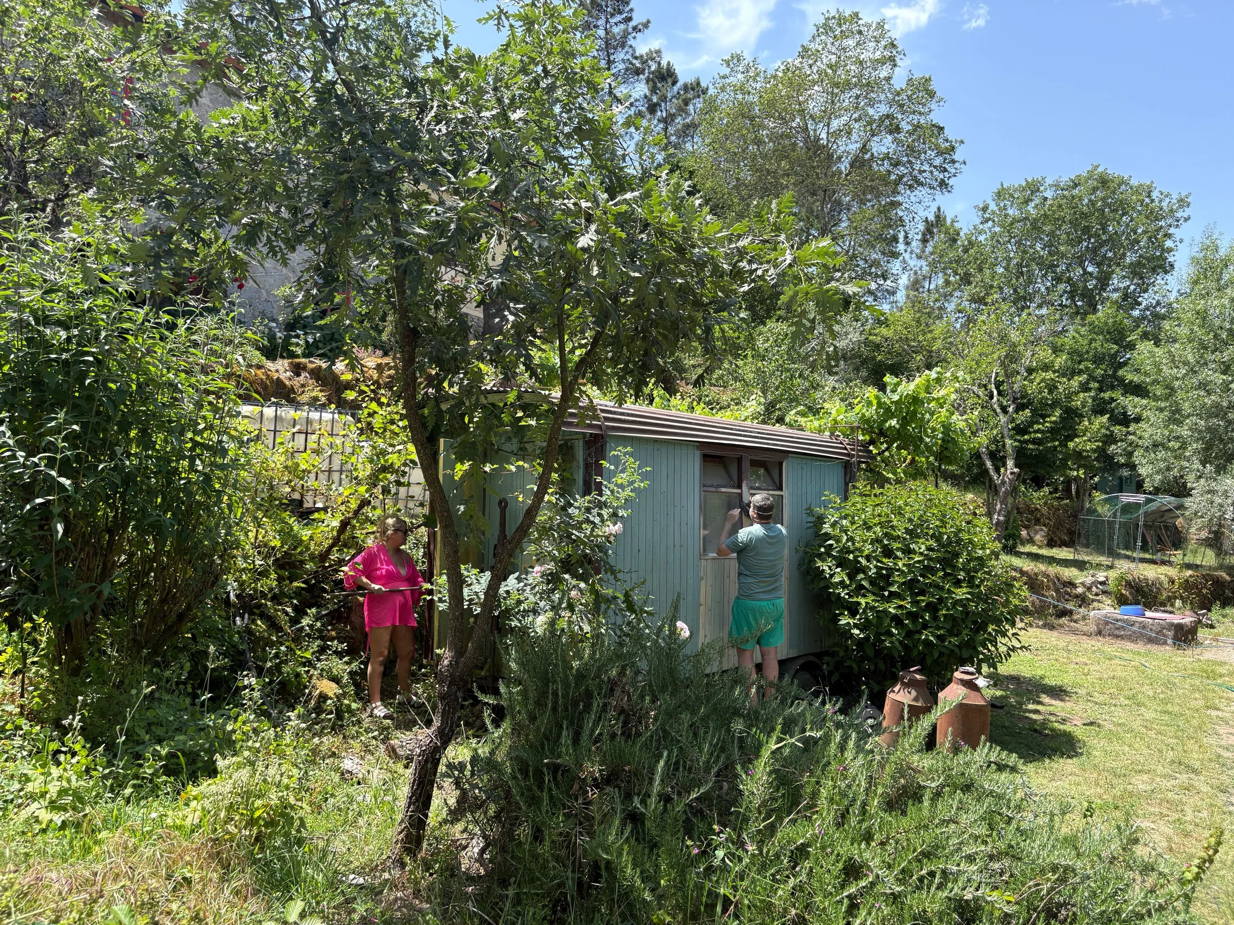Zwei Menschen stehen neben einem kleinen blauen Gartenhaus, umgeben von üppigem Grün und Bäumen bei sonnigem Himmel.