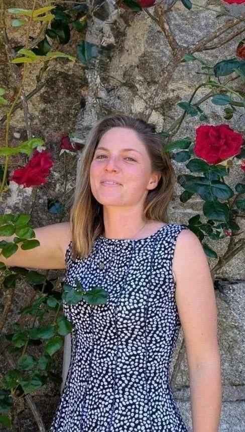 A woman with shoulder-length light brown hair in a sleeveless, black-and-white polka dot dress, standing in front of a stone wall with blooming red roses and green leaves.