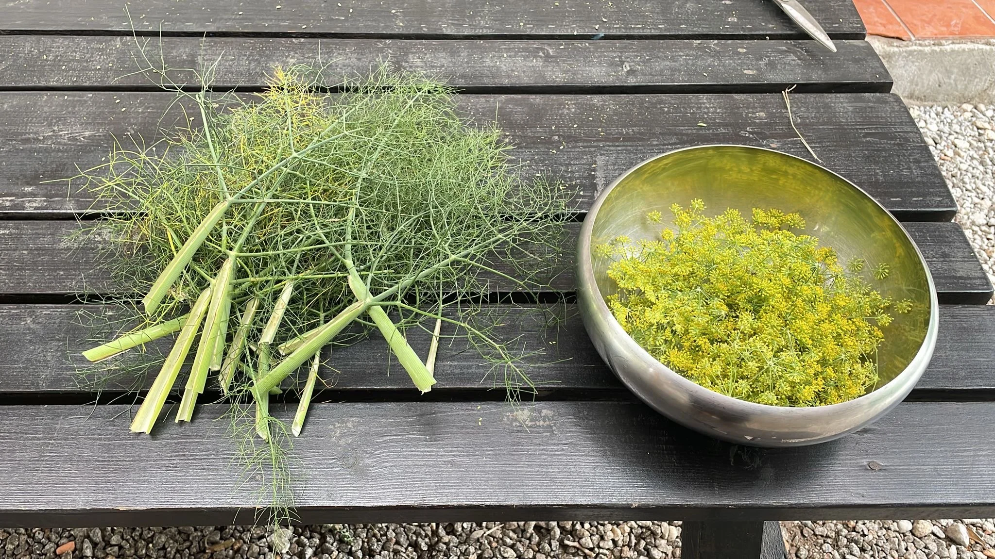 A black wooden outdoor table with fresh herbs on the left and a metal bowl of yellow flowering herbs on the right, with a concrete and gravel surface in the background.