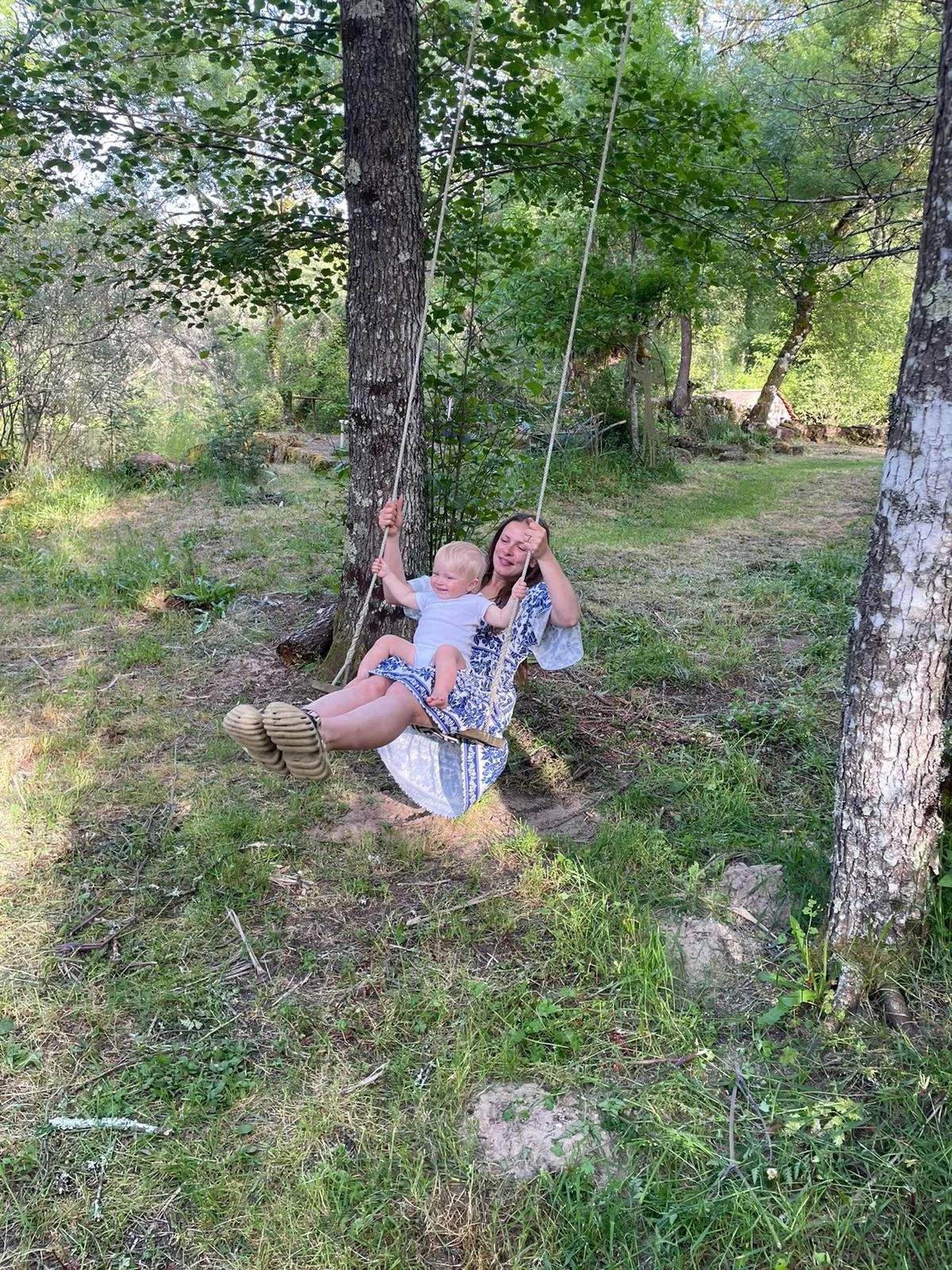 A woman and a young child are on a swing hanging from a tree in a grassy, wooded area. The woman is holding onto the swing's ropes while the child sits in her lap, both smiling and enjoying the moment. There are trees and greenery in the background.