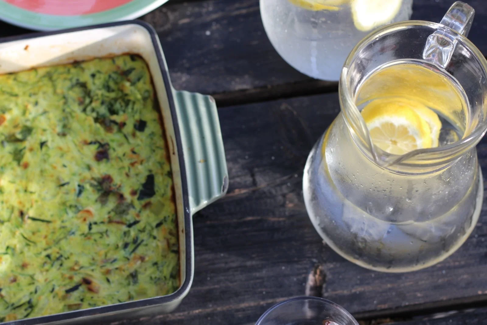 Close-up view of a rectangular dish of baked casserole, a clear glass pitcher filled with water and lemon slices, and a partially visible bowl of water with lemon slices on a dark wooden table.