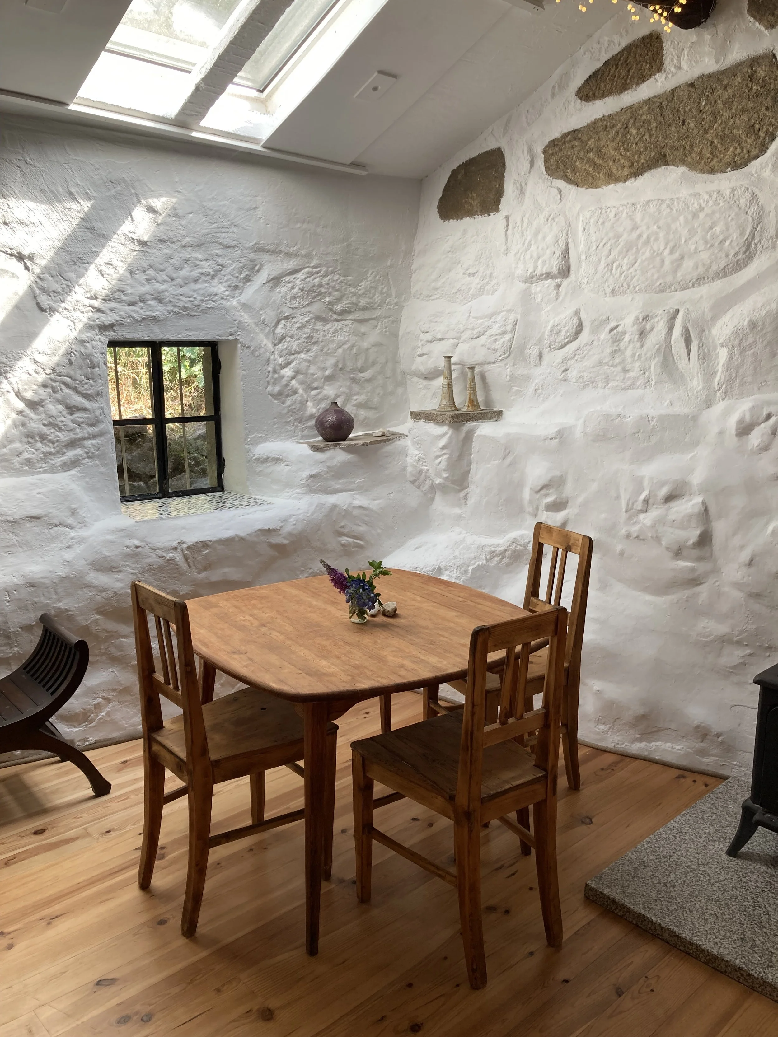 Ein Esszimmertisch aus Holz mit vier passenden Holzstühlen auf einem Holzboden. Die Wand ist weiß mit sichtbaren Steinen und eine kleine Fensteröffnung. Es stehen Dekorationsgegenstände auf einer Niesche, darunter Vasen. Die Decke hat ein Dachfenster