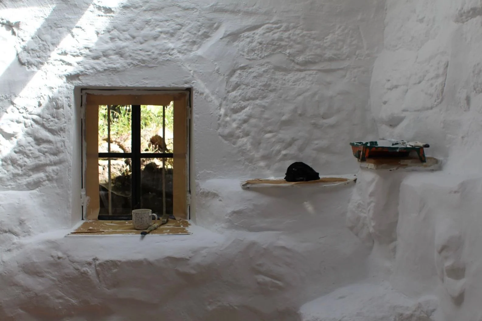 Innenraum mit weiß gestrichenen, unregelmäßig strukturierten Wänden, einem kleinen Fenster mit Gitter und zwei kleinen Holzregalen an der Wand. Auf dem Fensterbrett liegt eine Kaffeetasse und ein Werkzeug.
