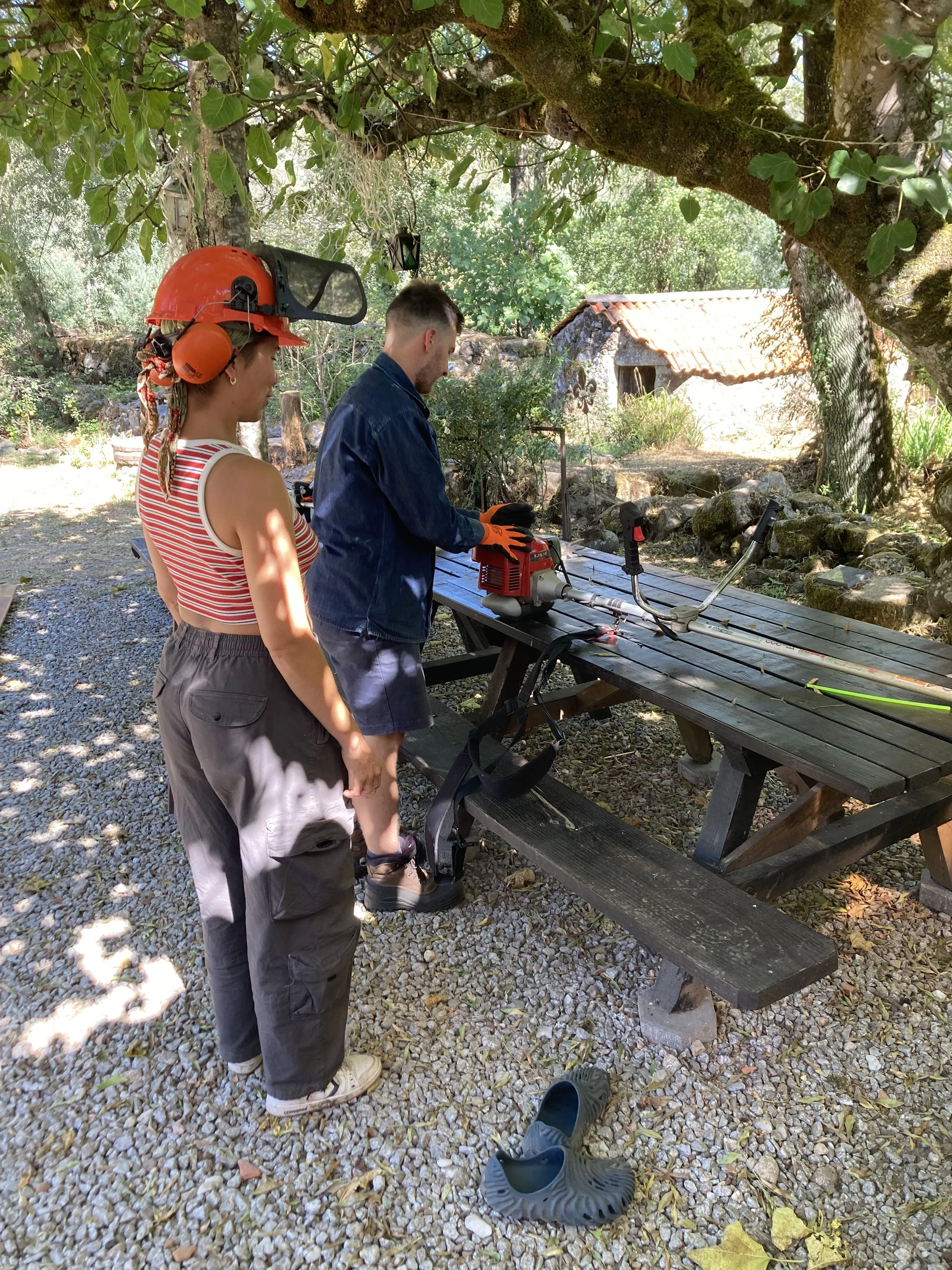 Two people stand outdoors under a large leafy tree, working with a piece of equipment on a wooden picnic table. One person wears a red helmet with attached hearing protection, and the other is dressed in a denim jacket and orange gloves. There are gardening tools, shoes, and a rock-lined ground surface.