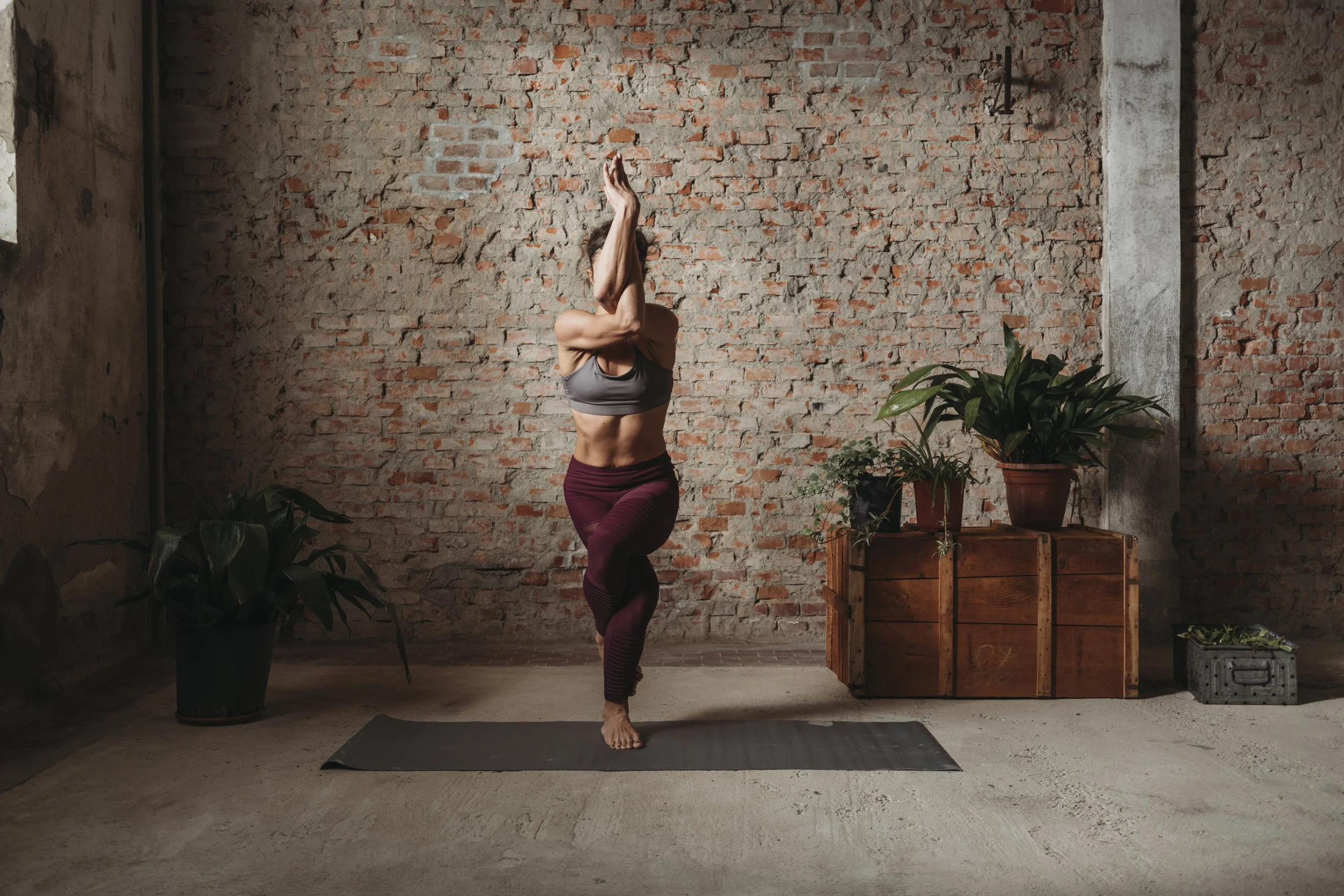 A woman practicing yoga in a rustic studio with a brick wall, plants, and wooden crates, standing on one leg with hands pressed together above her head.