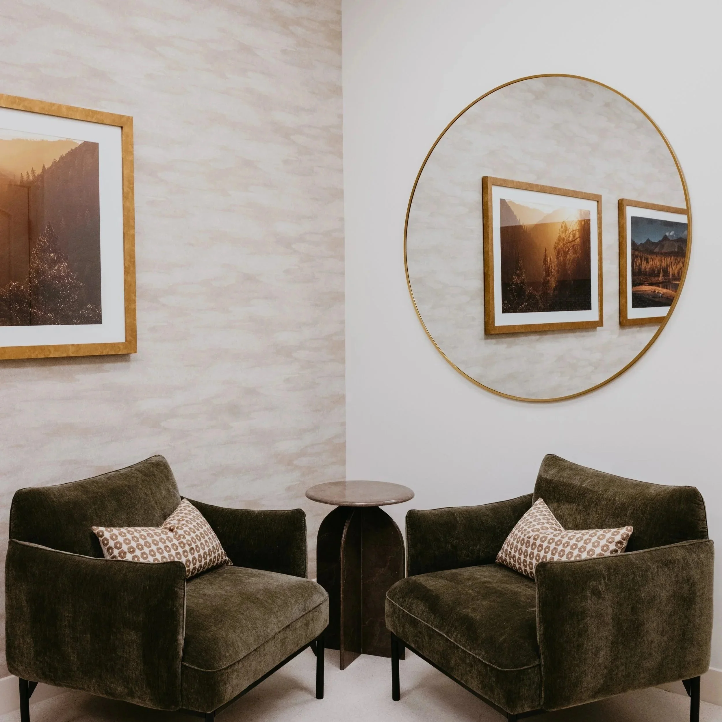 Living room with four dark gray armchairs arranged in a circle around a small round wooden table, beige pillows on the chairs, and two framed landscape photos on the wall, one large round mirror reflects the scene.