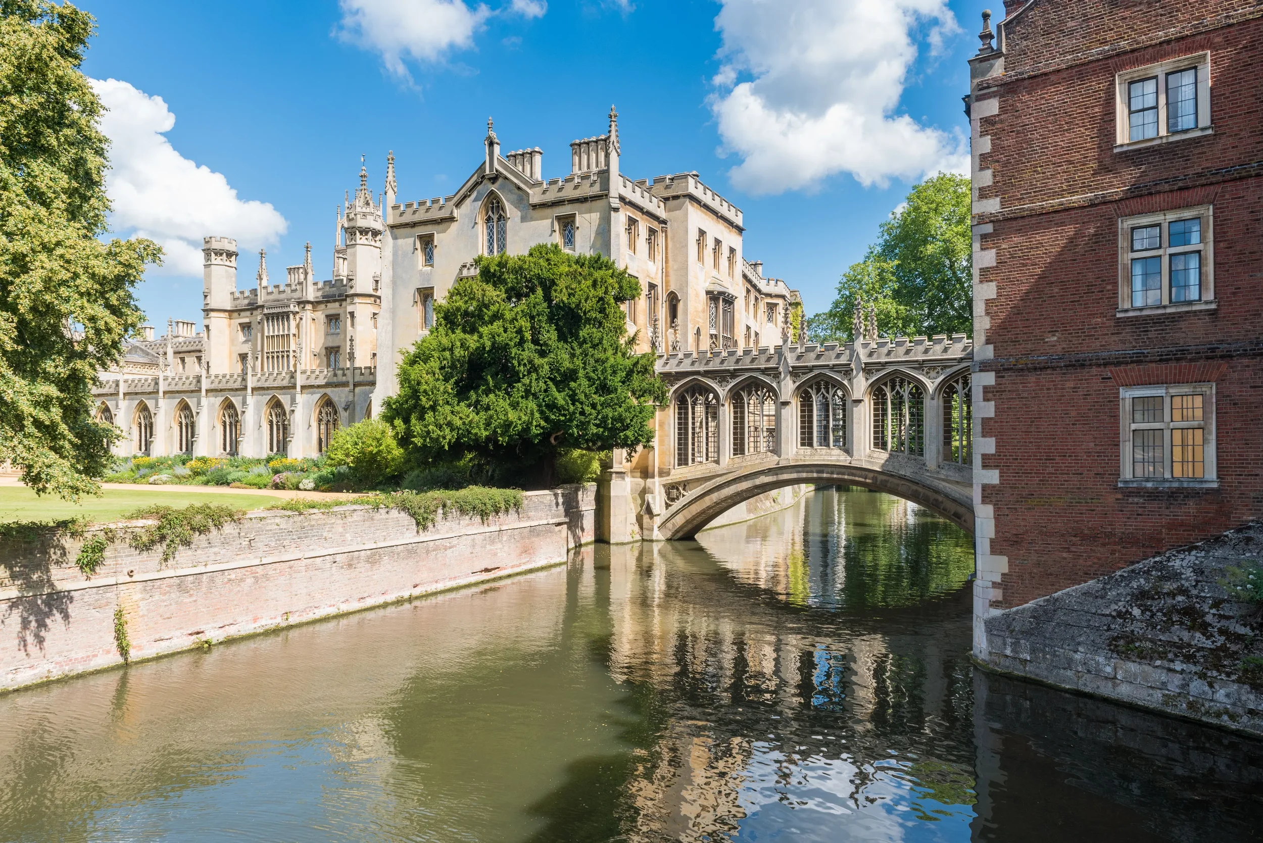 Historic castle with Gothic architecture beside a river, green trees, and a partly cloudy sky.