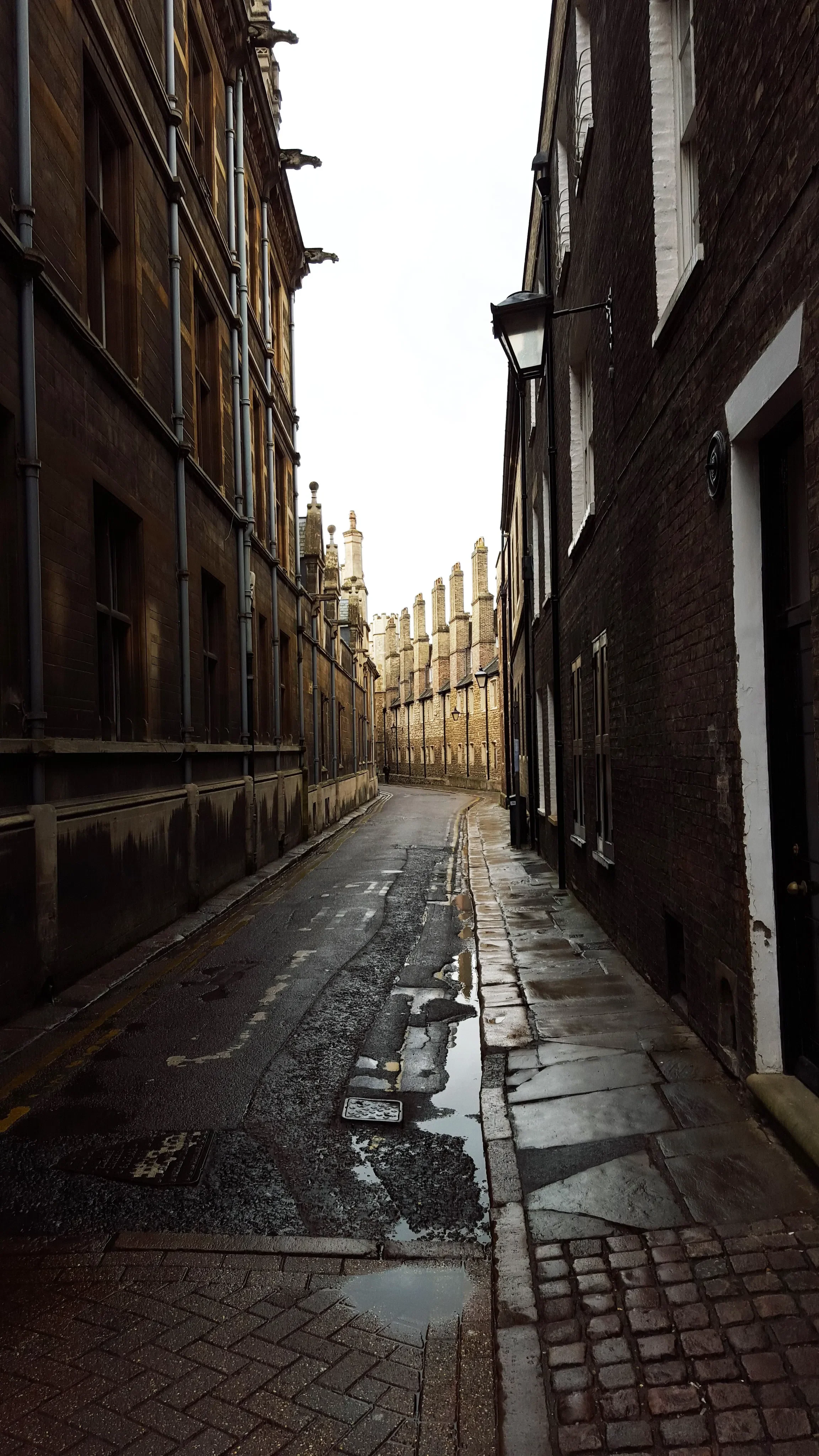 A narrow street with old brick buildings on both sides, wet pavement with puddles, and a cloudy sky overhead.