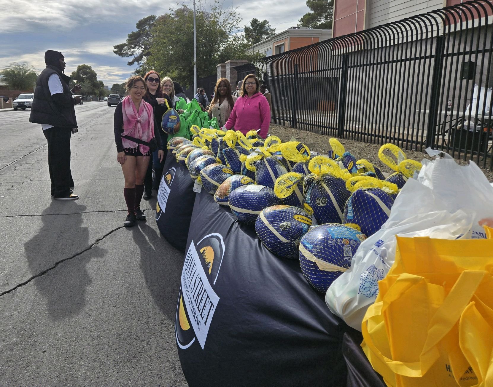 People standing behind tables filled with turkeys wrapped in blue netting, preparing for a charity or community event on a city street.