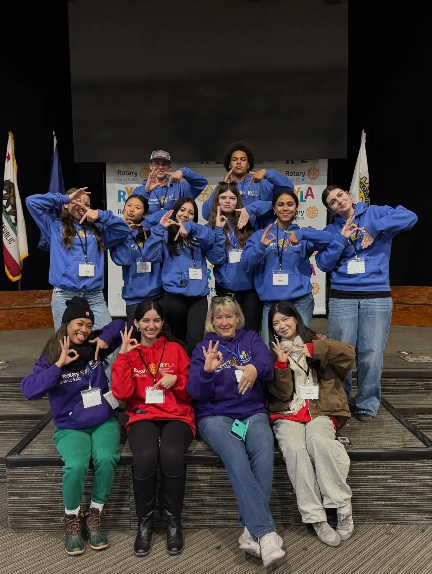 Group of young people and adults posing together on a stage, some making hand gestures and smiling, wearing colorful jackets and name tags at a Rotary event.