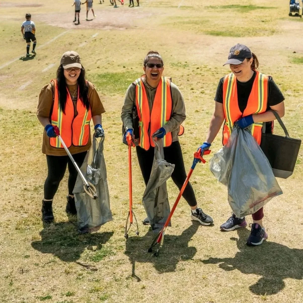 Three women wearing safety vests and gloves are holding trash pickers and bags, participating in a community cleanup on a grassy field, with children playing in the background.