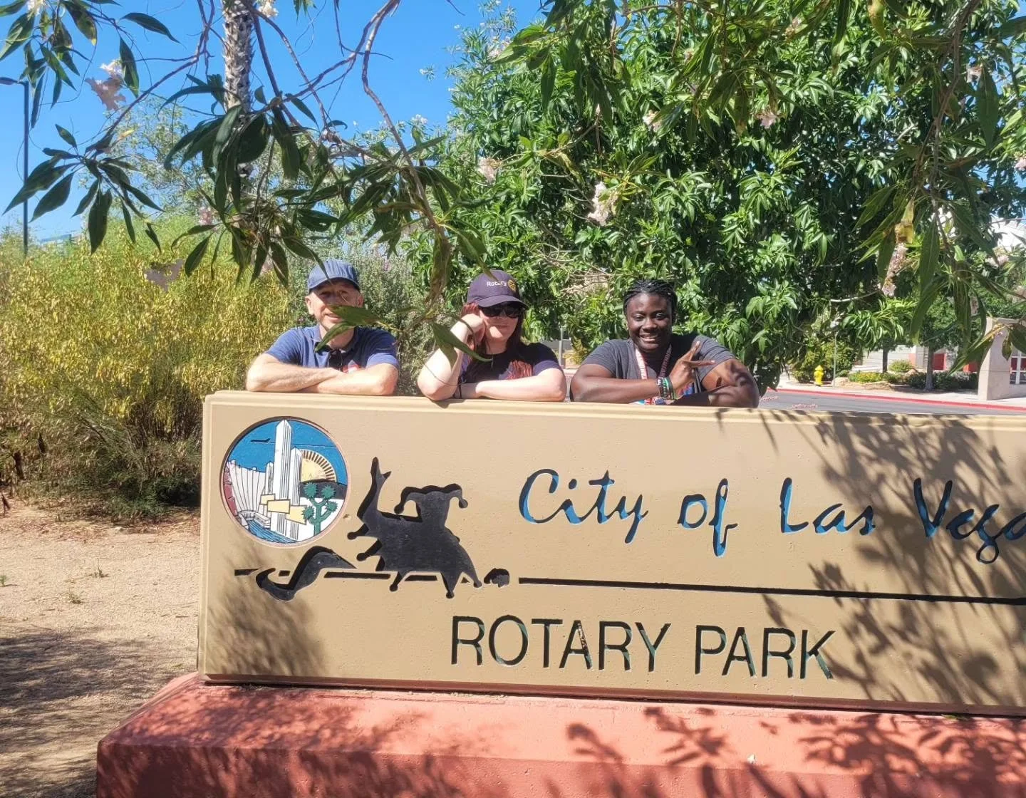 Three people sitting behind a sign that reads 'City of Las Vegas Rotary Park,' with trees and blue sky in the background.