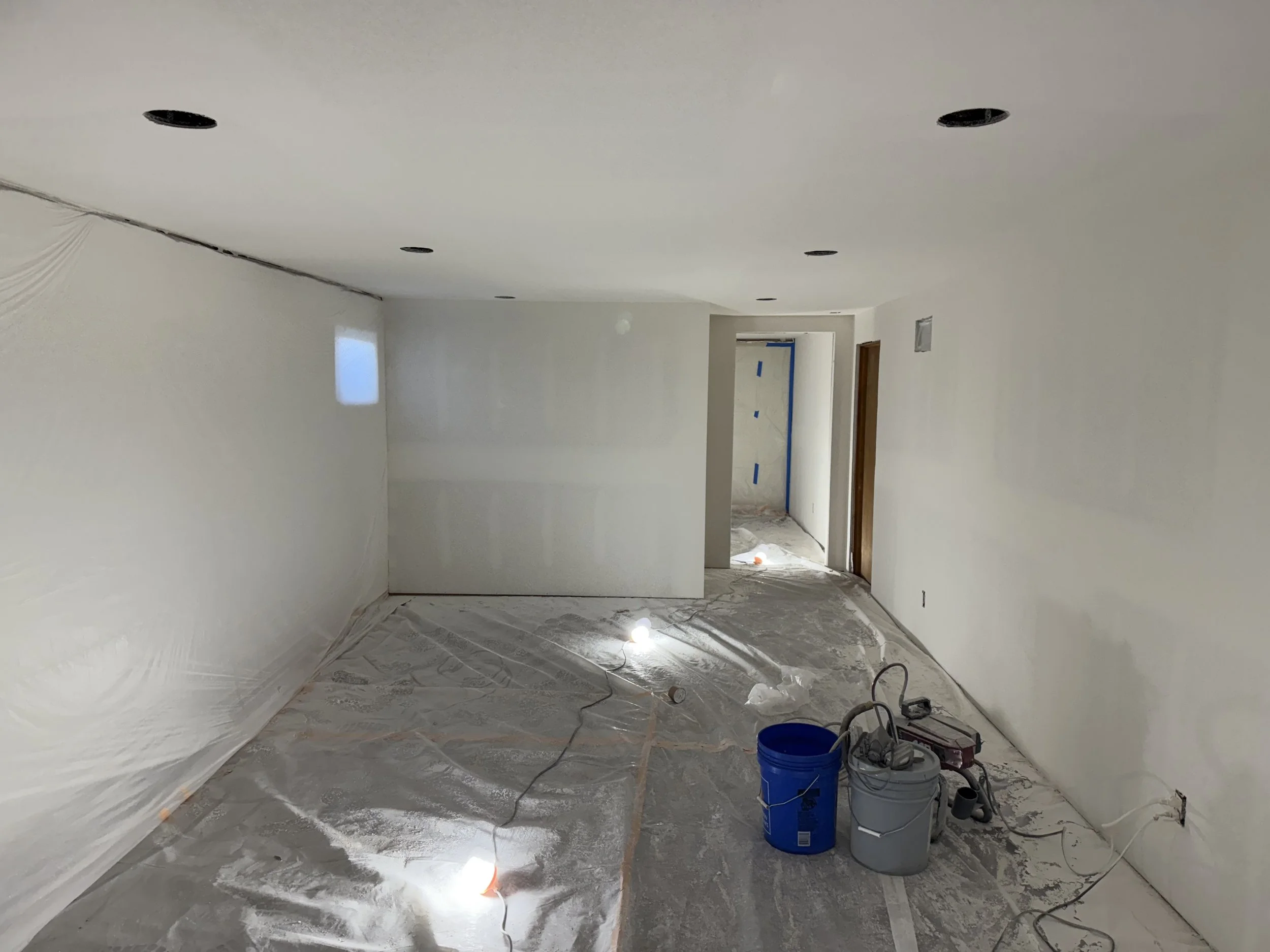 A room under construction with a white ceiling and walls, covered floors, and various construction tools and supplies including buckets, cords, and tools on the floor. Multiple ceiling holes for lighting are visible.