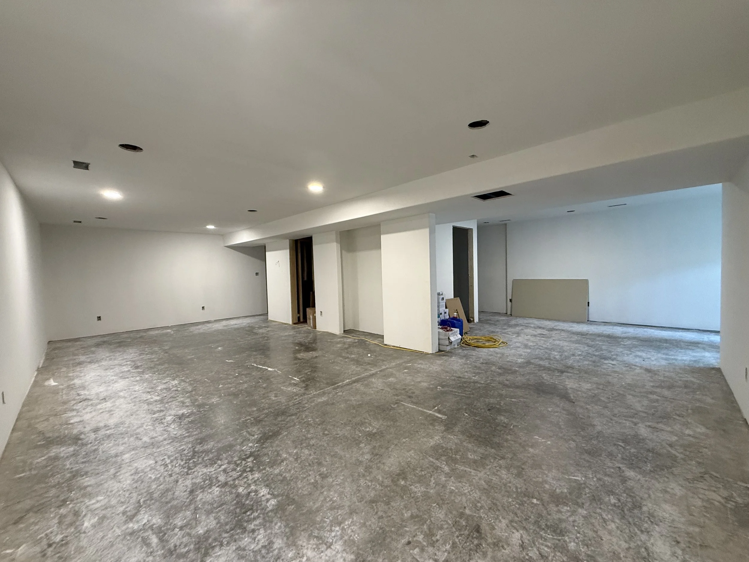 An unfinished room under construction with bare concrete floors, white walls, and visible ceiling fixtures without light bulbs.