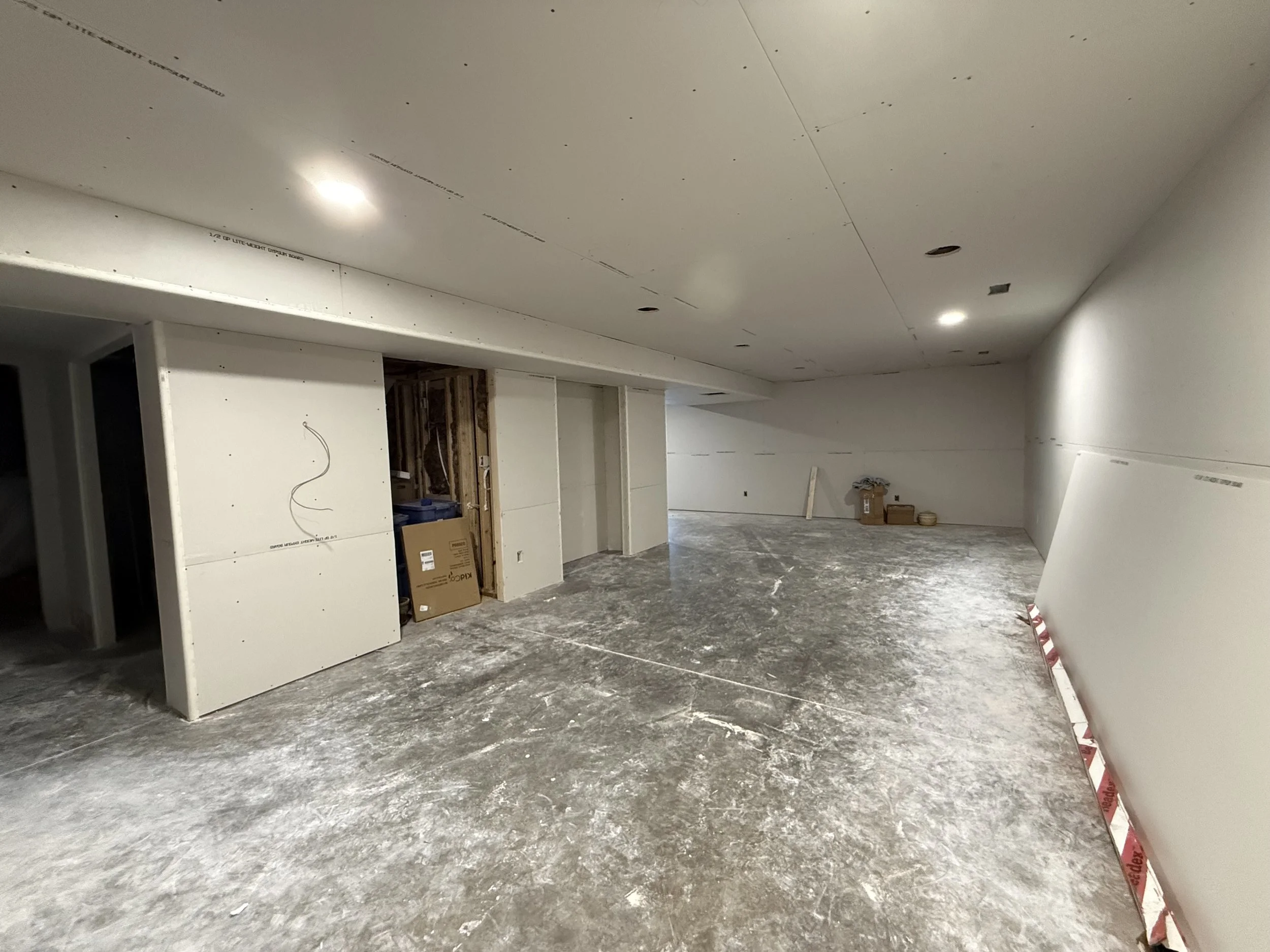 Interior of a building under construction with drywall installed on the ceiling and walls, unfinished flooring, and construction materials scattered around.