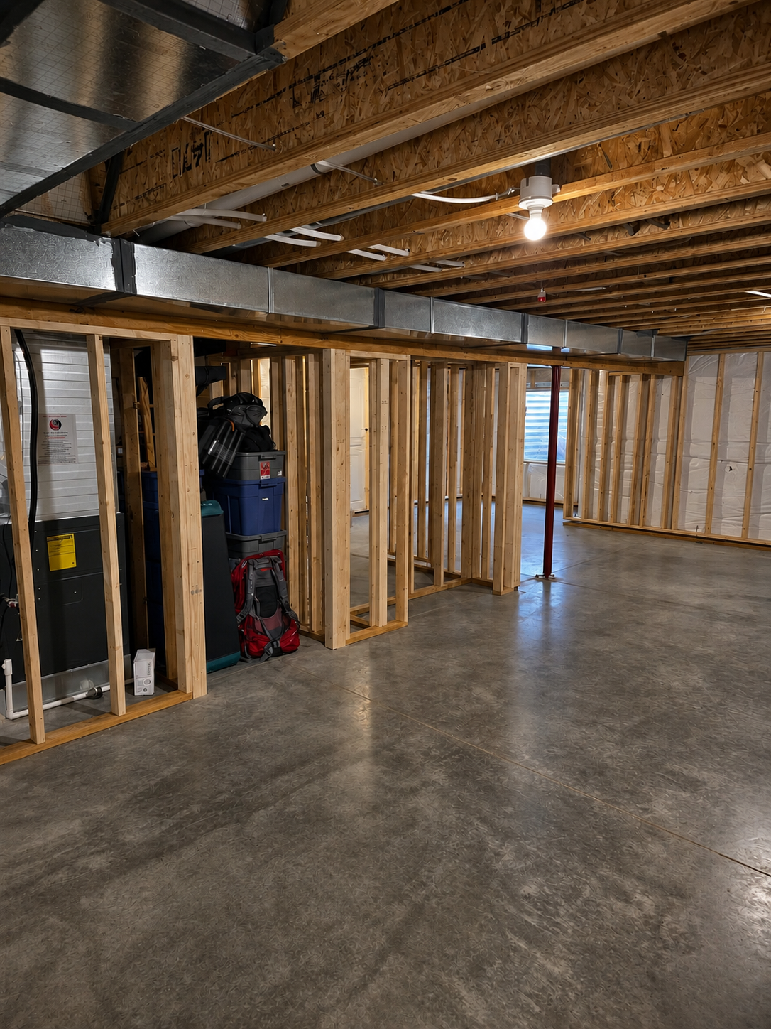 An unfinished room with exposed wooden framing, a gray concrete floor, and a visible HVAC duct running along the ceiling. There are tools and storage bins stacked against the wall.