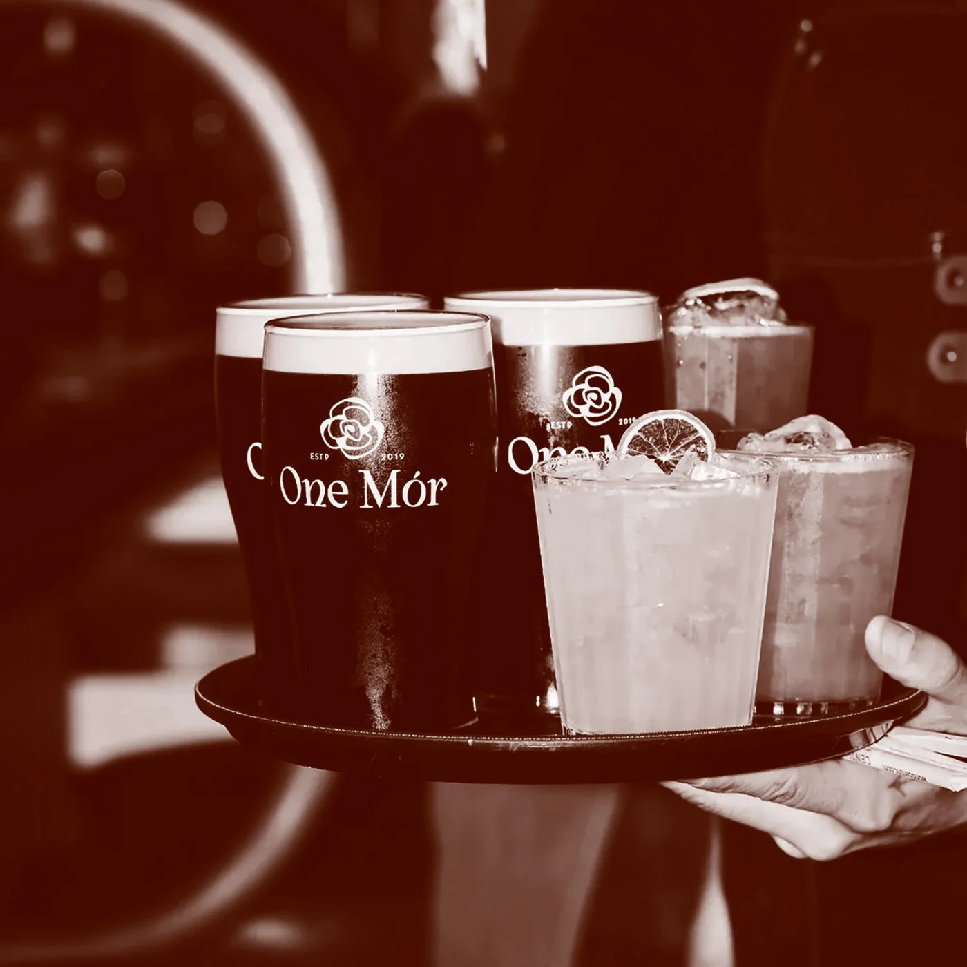 A waitress is holding a tray with four drinks: two glasses of dark beer with foam and 'One Mór' logos, and two glasses of cocktail with ice and lemon slices.