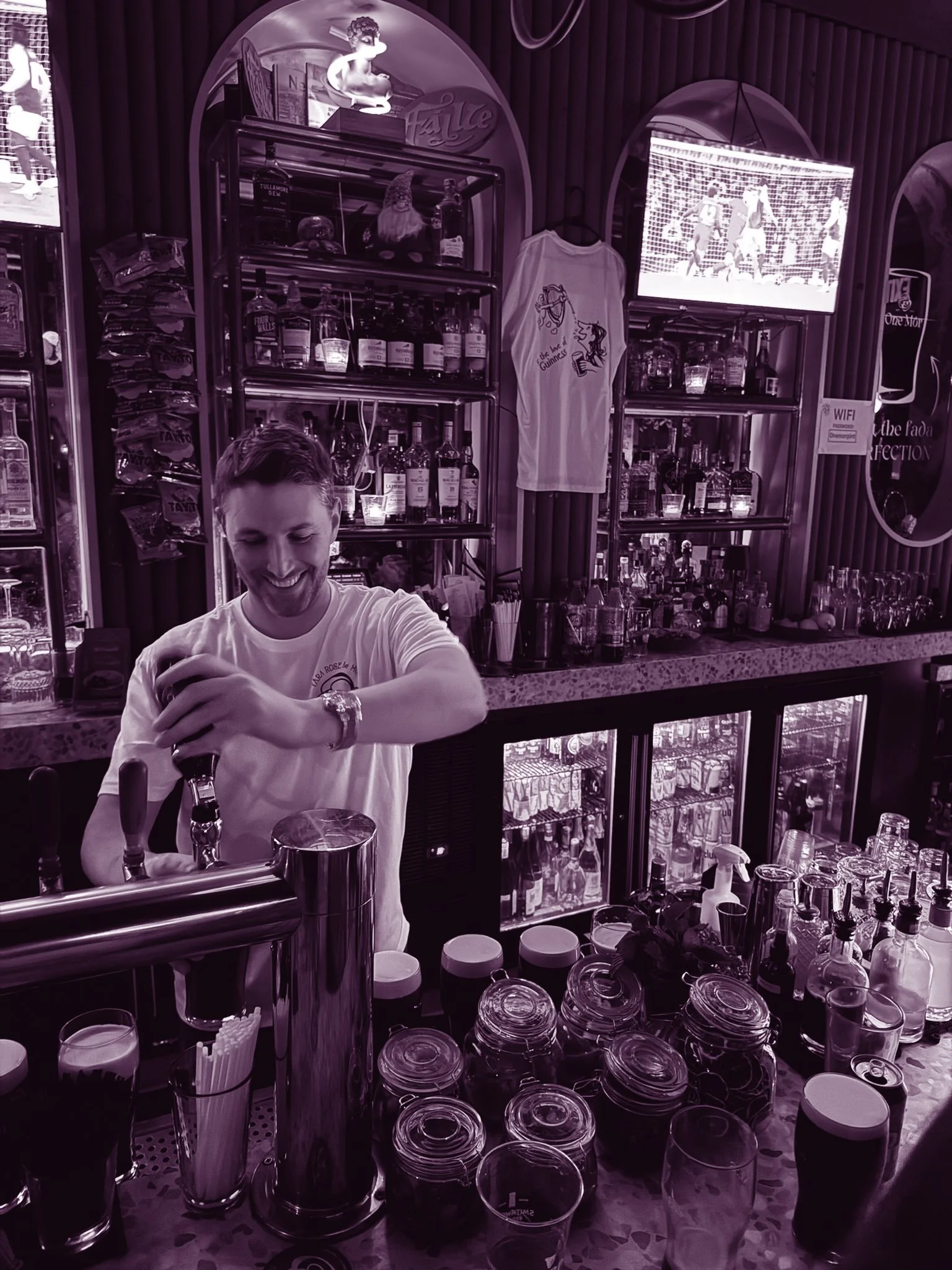 Bar counter with various bottles of alcohol, glasses, and bar tools, with a smiling bartender pouring a drink. Shelves behind the bar display bottles and decorations, and a television is mounted on the wall showing a sports game.