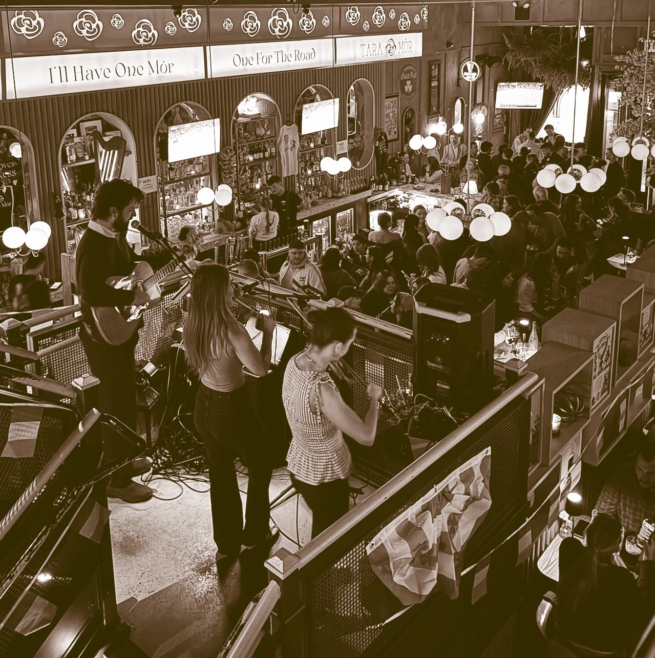 A crowded indoor venue with a live musical performance on a raised stage, featuring a guitarist and two female singers, surrounded by numerous audience members, decorative lighting, and a bar area in the background.