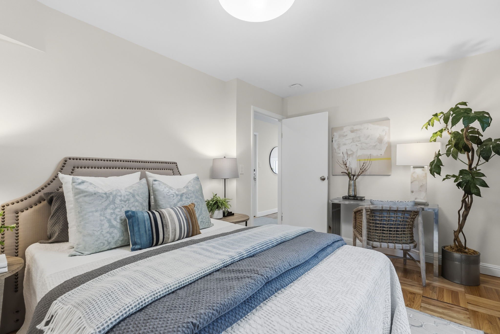 A cozy bedroom with a bed decorated with multiple pillows, a bedside lamp, a large potted plant, artwork on the wall, and a partially open door.
