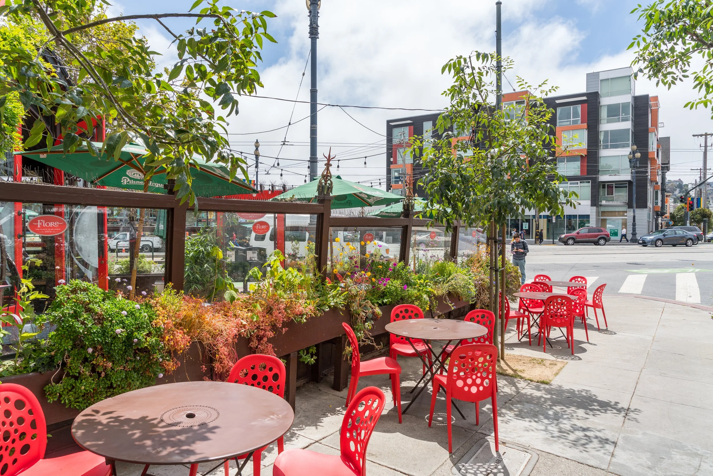 Outdoor patio with red seating and green umbrellas, surrounded by plants and flowers, in front of a city street with modern buildings and cars.