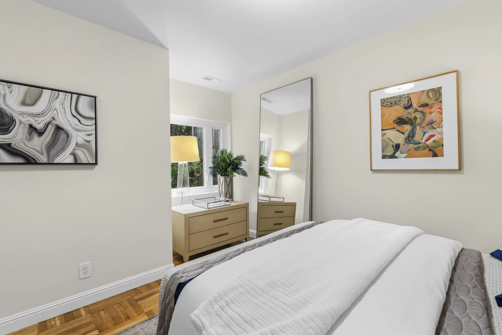 A neatly made bed with white bedding in a modern bedroom. Two light-colored wooden dressers are positioned on either side of a large mirror. Two table lamps and a potted plant are on the dresser near the mirror. Abstract artwork hangs on the walls, a