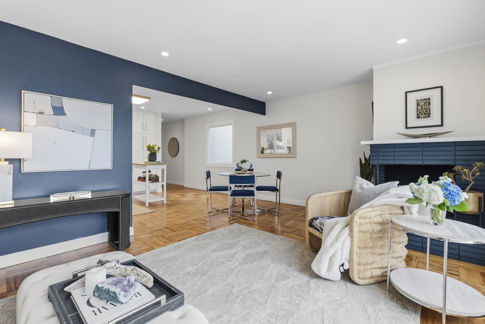 A modern living room with a blue accent wall, white ceiling and walls, hardwood floors, and ceiling lights. There is artwork on the walls, a dining table with chairs, a fireplace with a decorative object on the mantel, and various plants and flowers.