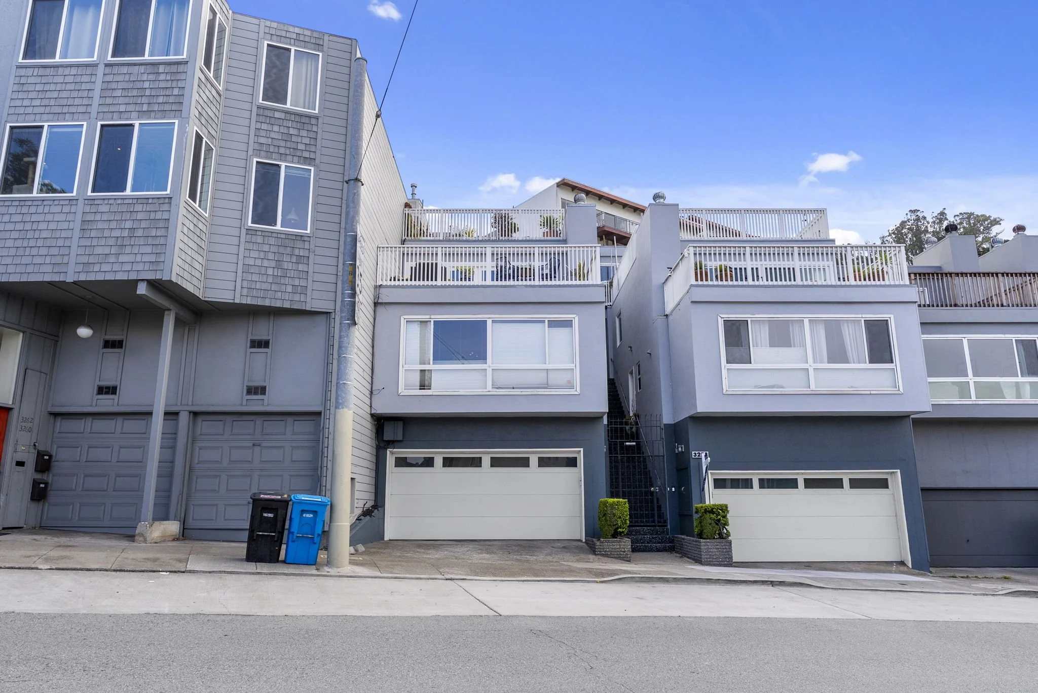 Front view of a multi-story residential building with gray exterior, large windows, balconies with plants, and a garage with two trash bins outside on the street under a clear blue sky.