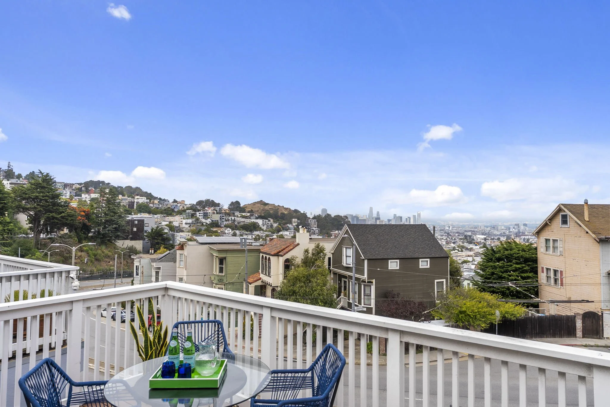 View from a balcony overlooking a hilly neighborhood with houses, trees, and a city skyline in the distance under a partly cloudy blue sky.