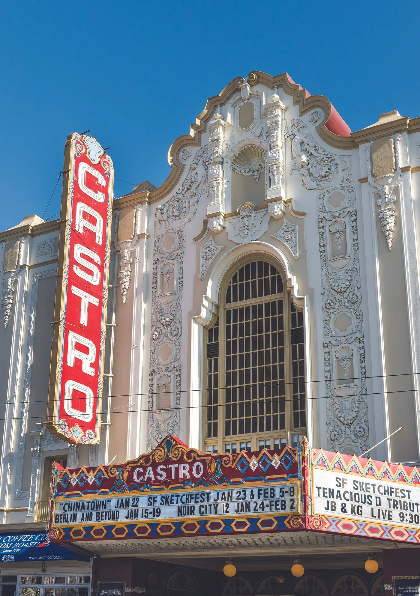 The Castro Theatre, an ornate white historic building with detailed decorative carvings and a large red vertical sign reading 'CASTRO' on the front. The marquee lists upcoming events and dates, with bright, colorful border decorations at the entrance