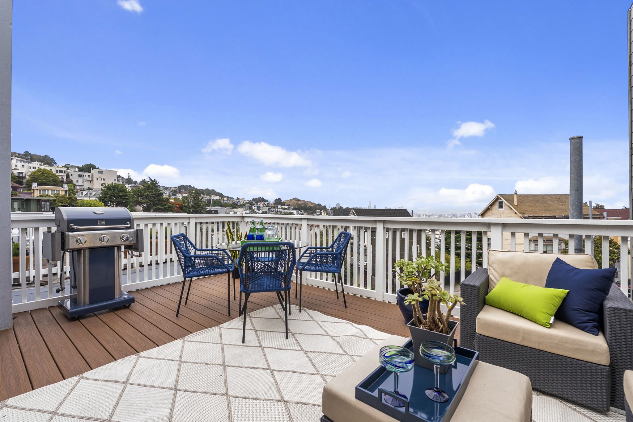A balcony with outdoor furniture, including a small dining table with blue chairs, a gray sofa with colorful pillows, a potted plant, and a barbecue grill, overlooking a neighborhood with houses and trees under a blue sky with clouds.