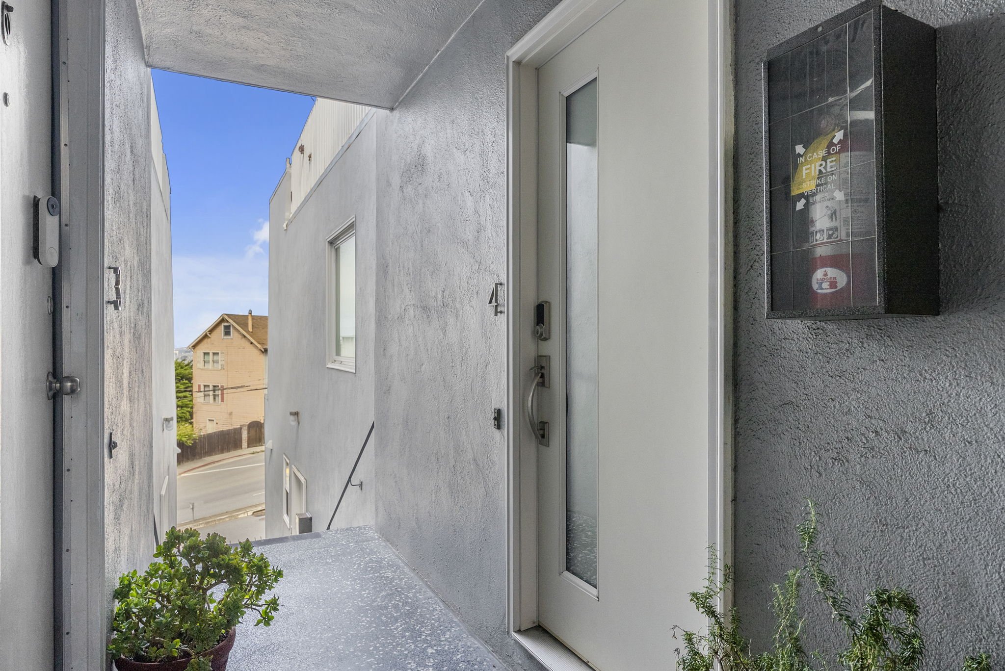 Apartment building entrance with white door, potted plants, fire extinguisher box, and clear sky above.