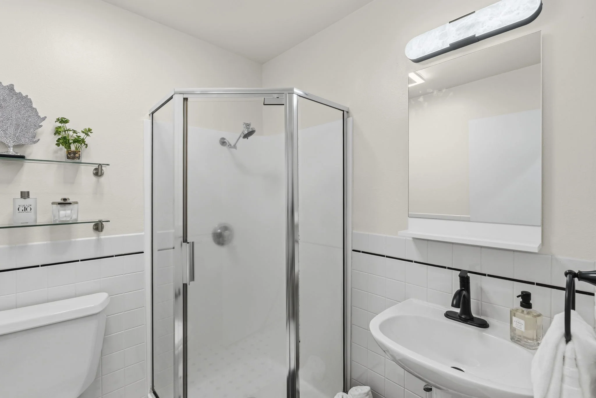 A small bathroom with a corner glass shower stall, white sink with black faucet, round mirror, window, and minimal decor including a plant, perfume, and soap bottles.