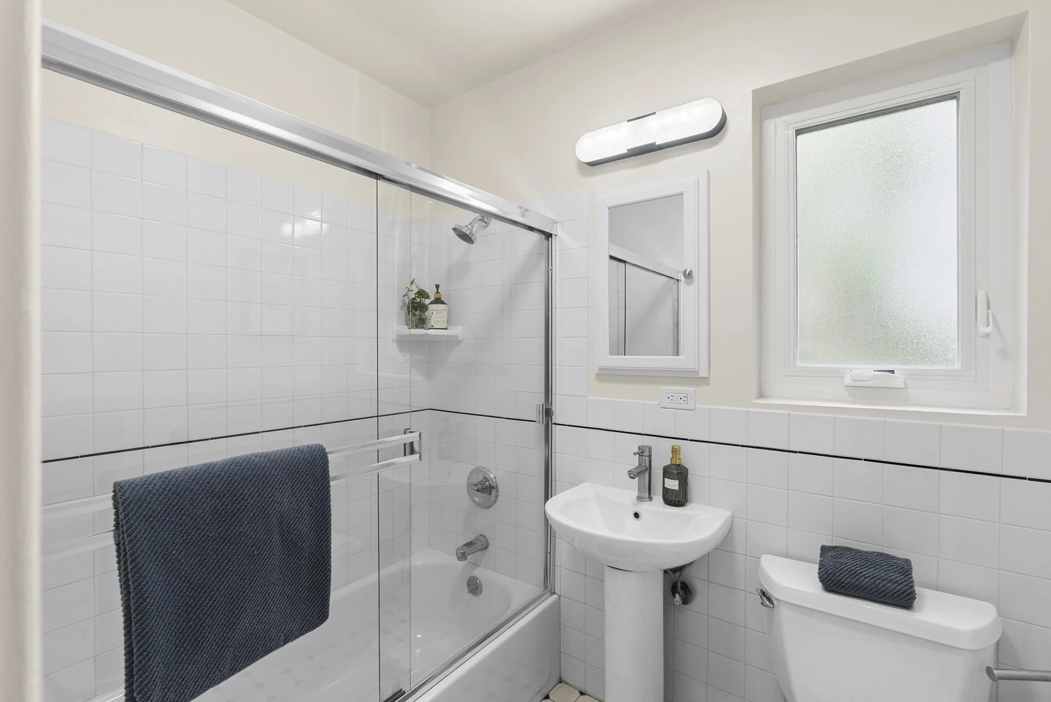 Bathroom with white tiled walls, a glass shower enclosure, a small sink, a toilet, a window with frosted glass, and a light fixture above the mirror.