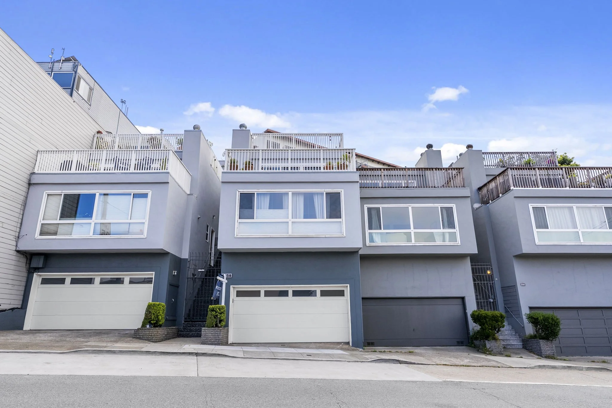 Three-story modern gray residential building with large rectangular windows, balconies with white railings, and a garage on the ground level. There are small landscaped bushes in front and a clear blue sky above.