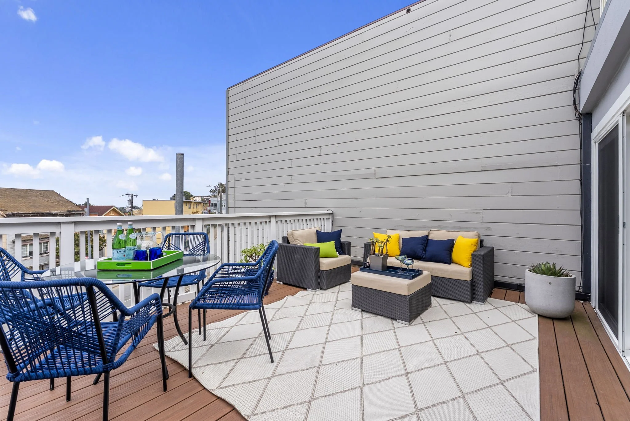 An outdoor balcony with a table and chairs, a seating area with cushions, potted plants, and a view of neighboring buildings and blue sky.
