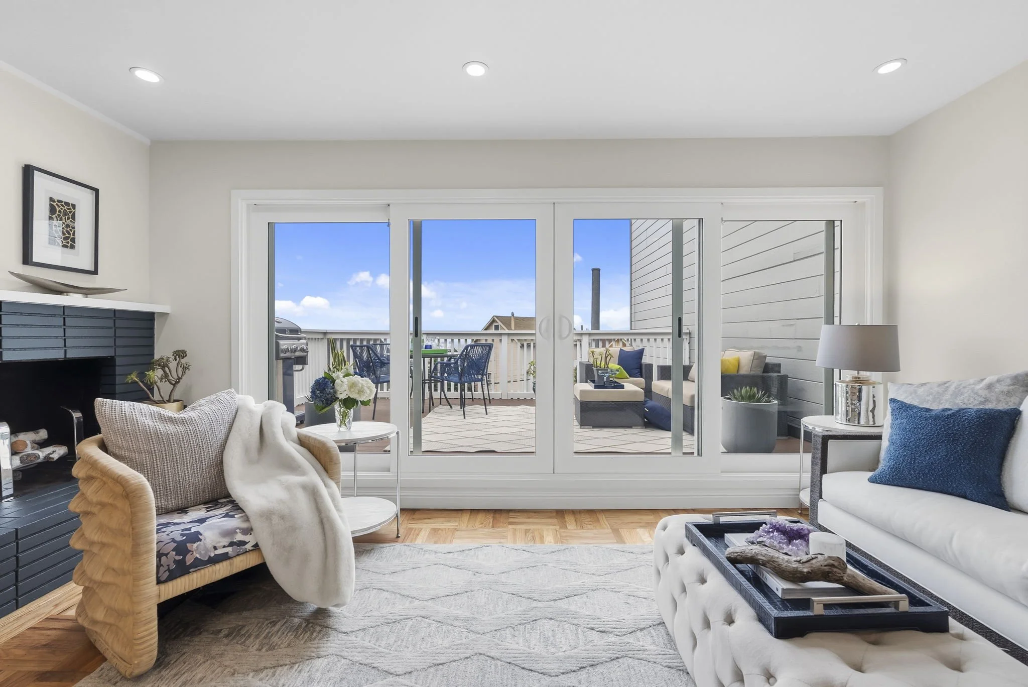 Living room with sliding glass doors leading to an outdoor patio with seating, a grill, and a blue sky.