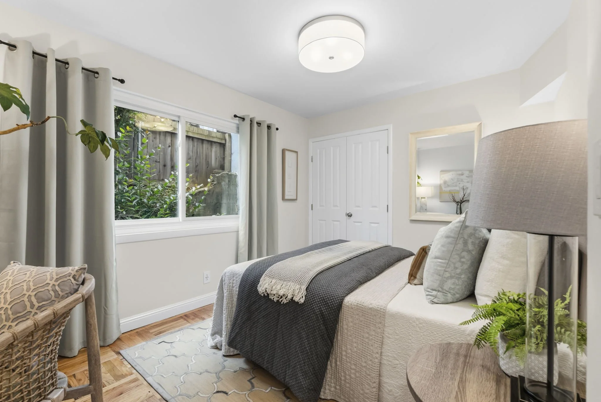 A bedroom with a bed, pillows, and a blanket, featuring large windows with curtains, a mirror, and houseplants, illuminated by a ceiling light.
