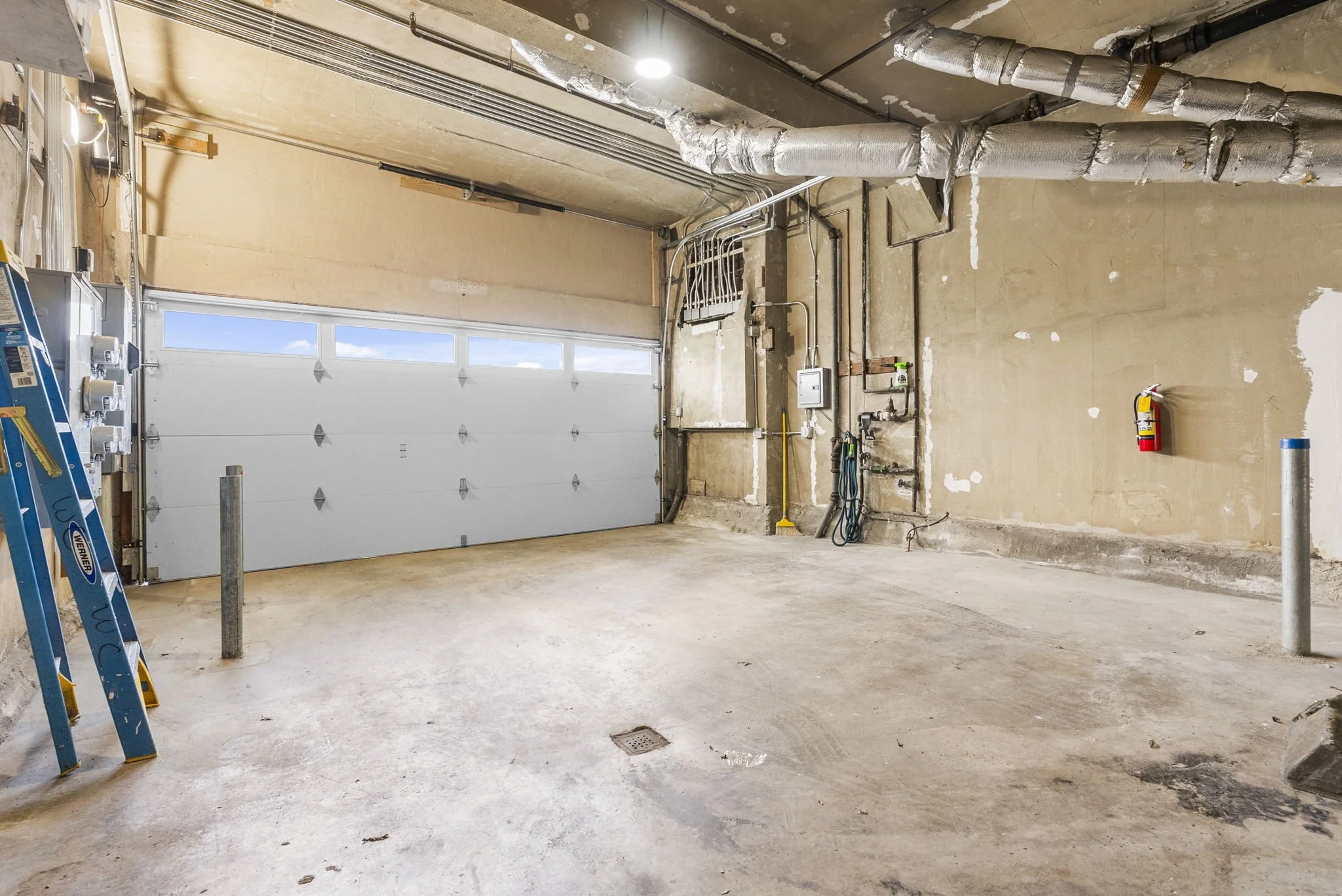 Empty garage with an unfinished interior, exposed pipes and wiring, a ladder on the left side, and a fire extinguisher on the right wall.