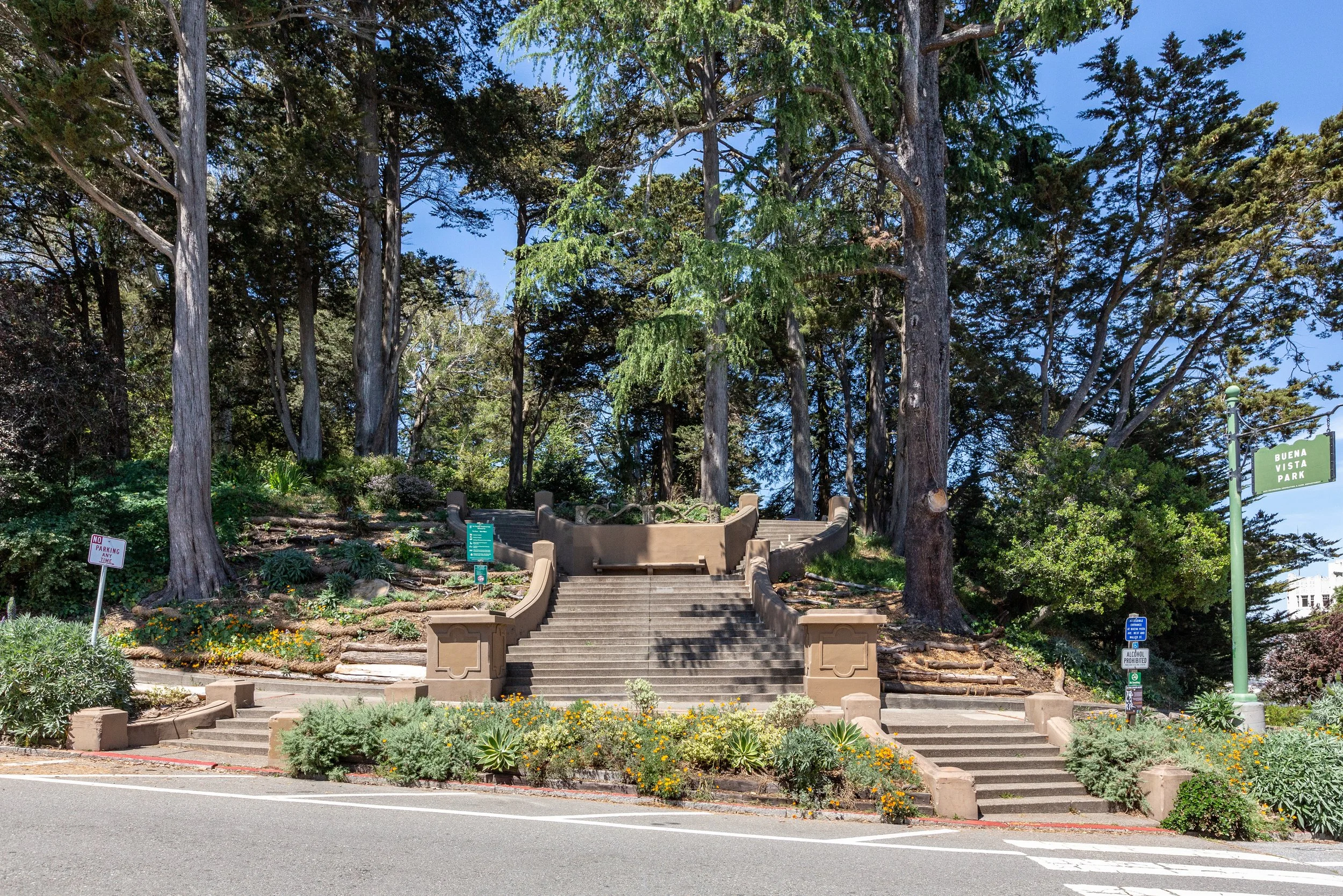 Stone staircase leading up to a park area with large trees, shrubs, and flowers, with signs indicating park rules.