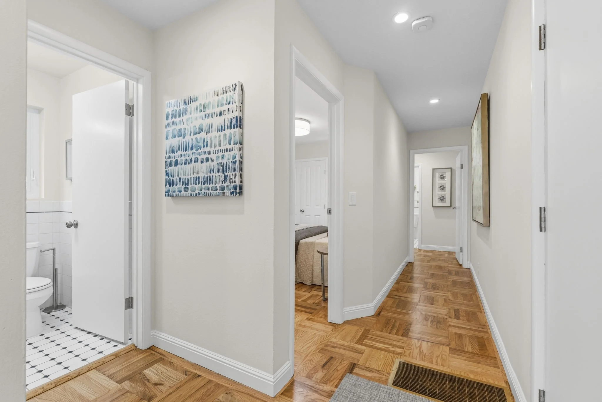 Interior view of a hallway with hardwood floors, white walls, and a ceiling with recessed lighting. Artworks hang on the walls, and multiple doorways lead to different rooms, including a bathroom with a white toilet visible through one door.